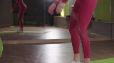 Beden eğitimi - Pembe spor kostümlü genç bir kadın ayna fitness salonunda çömeliyor.