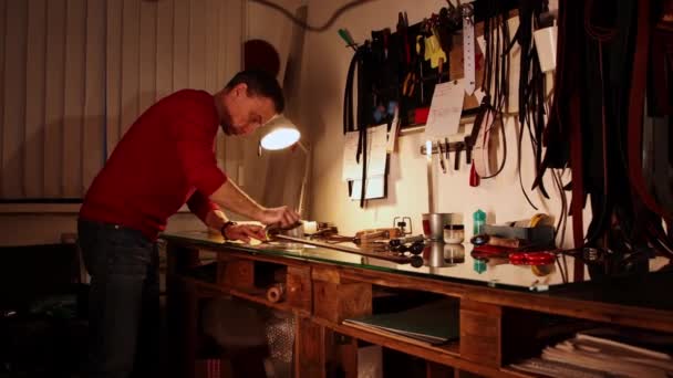 Taller de cuero: un hombre trabaja con un cinturón en blanco en la tenue luz de la lámpara — Vídeos de Stock