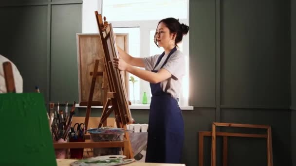 Art studio - young woman puts a canvas on an easel and starts painting — Stock Video