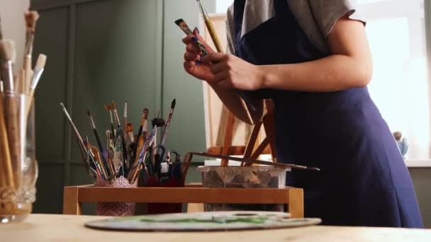 Художня студія - молода жінка художник підбирає пензлі з банок — стокове відео