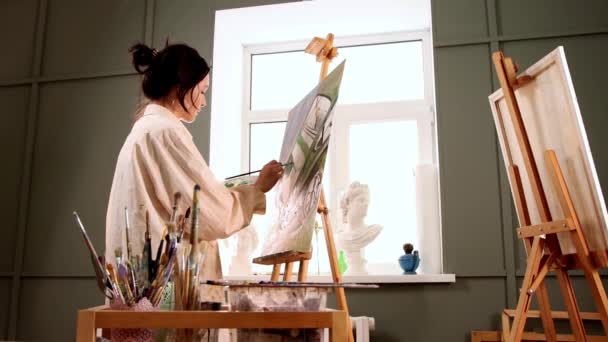 Estudio de arte - mujer joven con camisa blanca manchada cambiando pinceles y dibujando una pintura — Vídeos de Stock