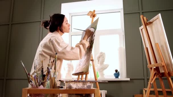 Kunstatelier - junge hübsche Frau im fleckigen weißen Hemd wechselt die Pinsel und zeichnet ein Gemälde — Stockvideo