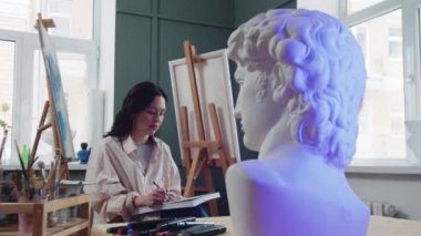 Bir kadın sanatçı, bir sanat stüdyosundaki mermer büstünden eskizler çiziyor.