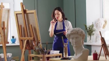 Resim stüdyosunda resim çizen bir kadın ressam saçını bir sehpanın önüne bağlıyor.