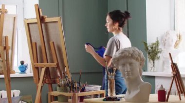 Bir kadın sanatçı bir sanat stüdyosuna girer ve üzerine önlük bağlar.