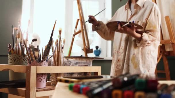 Art studio - young woman artist in white color stained shirt mixing colors on palette and drawing a painting — Stock Video