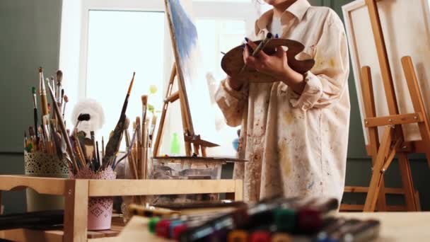 Art studio - giovane e felice artista donna in camicia colorata di colore bianco che mescola i colori sulla tavolozza e disegnare un dipinto — Video Stock