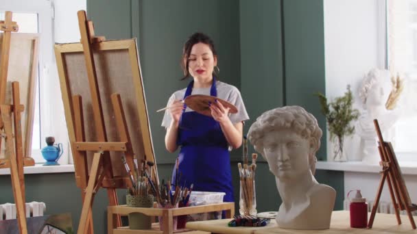 Donna carina e sorridente lavora con un dipinto su cavalletto nello studio d'arte — Video Stock