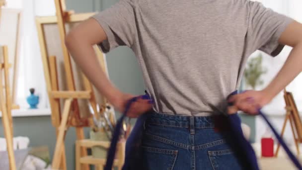 Uma mulher artista entra em um estúdio de arte e amarra um avental — Vídeo de Stock