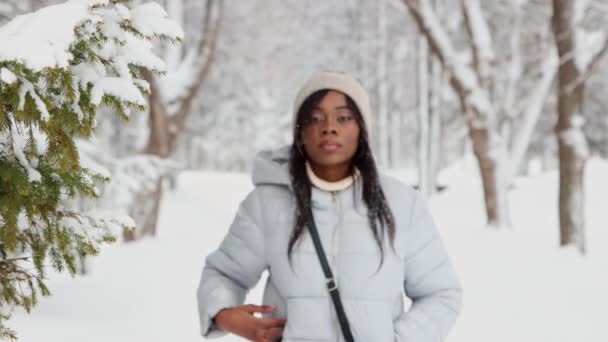 Молодая черная женщина в белой куртке гуляет по зимнему лесу — стоковое видео