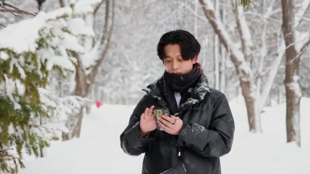 젊은 아시아인 남자가 겨울 숲에서 핸드폰을 사용하고 친구를 만나기 위해 걸어 나간다 — 비디오