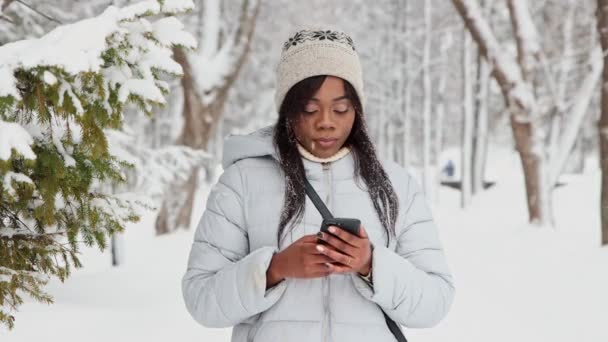 Молодая черная женщина в белой куртке пользуется телефоном в зимнем лесу — стоковое видео