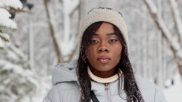 Молодая черная женщина в белой куртке стоит в зимнем лесу и улыбается в камеру — стоковое видео