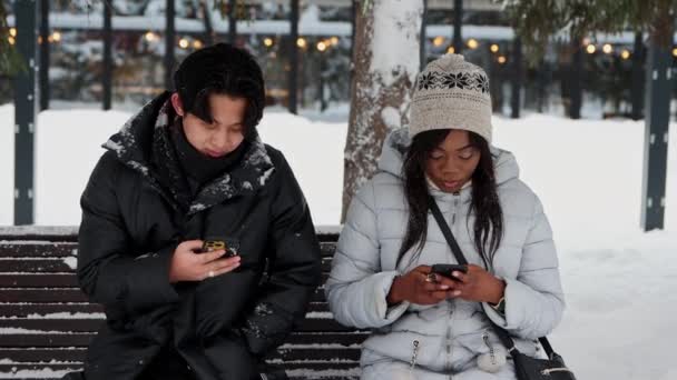 Mladý asijský muž a mladá černoška sedí na lavičce v zimním parku a dívá se na jejich telefony — Stock video