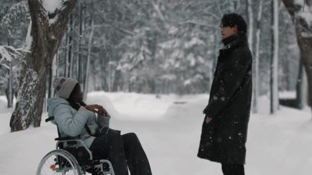 Joven negro mujer en un silla de ruedas hablando con su asiático macho amigo en invierno parque — Vídeos de Stock