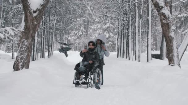 Молодая пара азиатских парней в инвалидной коляске и его черная подружка делают селфи в зимнем лесу — стоковое видео