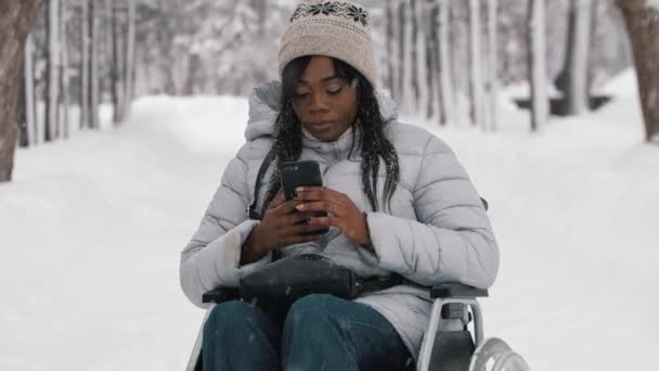 Молодая черная женщина сидит в инвалидной коляске и пользуется телефоном в зимнем лесу — стоковое видео