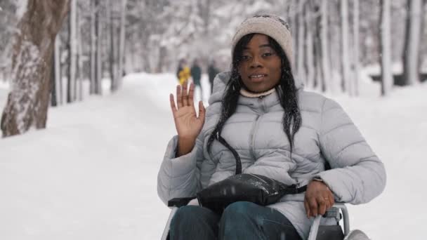 Молодая черная женщина сидит в инвалидной коляске в зимнем лесу и машет рукой — стоковое видео