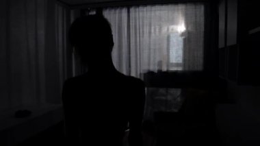 Spor elbiseli genç bir kadın perdeleri açıyor ve odaya ışık giriyor.