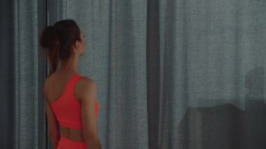 Spor elbiseli genç ve güzel Silim kadın perdeleri açar ve odaya ışık tutar.