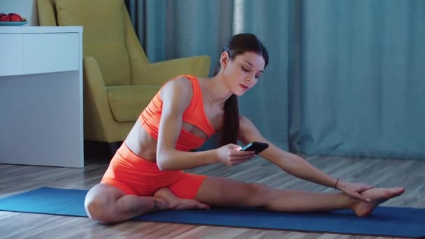 Junge hübsche Frau in orangefarbener Kleidung macht Stretching und schaut auf ihr Handy — Stockvideo