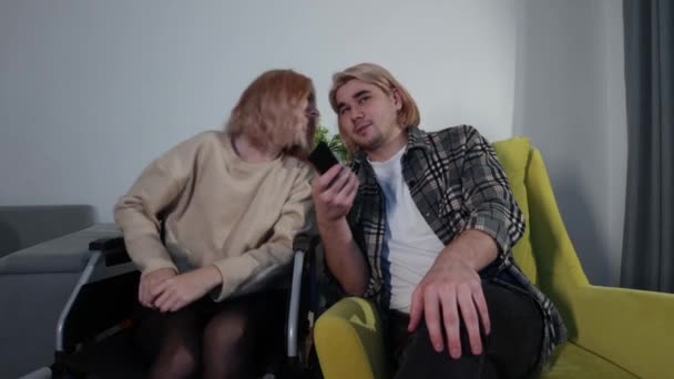 Νεαρός άνδρας και γυναίκα σε αναπηρική καρέκλα κάθονται στο σαλόνι — Αρχείο Βίντεο