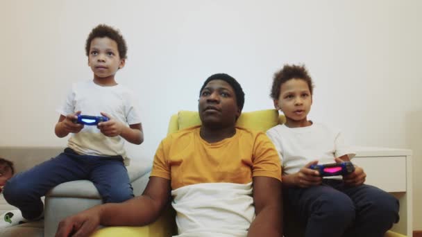 Dois meninos negros jogando um jogo usando joysticks e seu pai se senta entre eles e ajudando — Vídeo de Stock