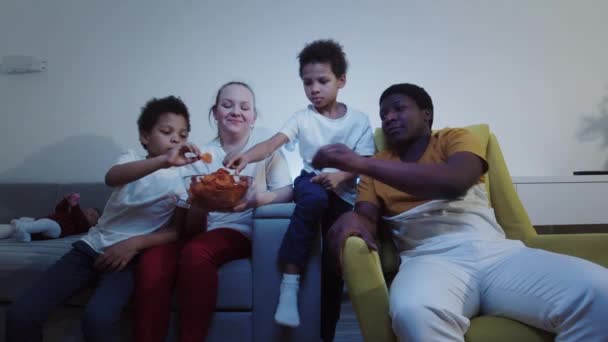 Multirace familia viendo la televisión y comiendo bocadillos por la noche — Vídeos de Stock