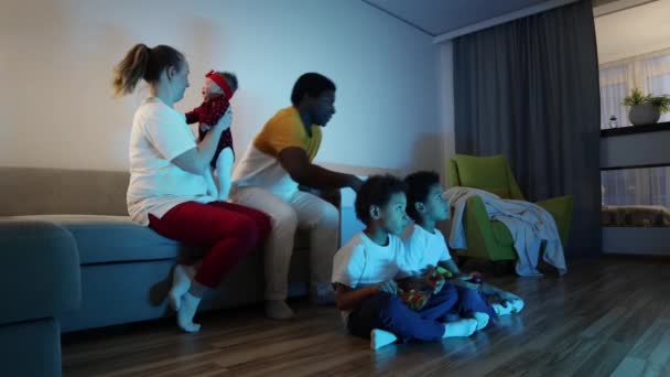 Çok kültürlü bir aile odada televizyon izler. Baba ikiz oğullarını battaniyeyle örter. — Stok video
