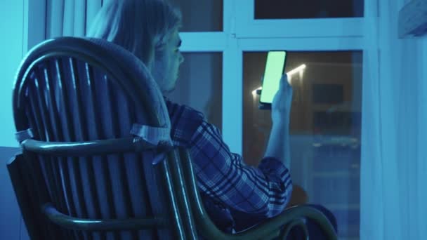 Young blonde man sitting in a rocking chair and looking at his phone in blue lighting — Stock Video
