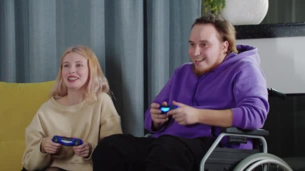 Feliz casal de mulher loira e homem em uma cadeira de rodas jogando jogo usando joysticks — Vídeo de Stock