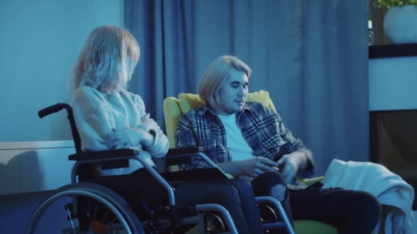 Ung man och kvinna i rullstol tittar på TV och mannen som täcker hennes ben med en filt — Stockvideo