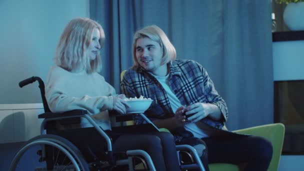 Young man and woman in a wheelchair watching TV - eating popcorn and drinking soda — Stock Video