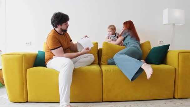 Família sentada no sofá amarelo e pai segurando um caderno com desenhos — Vídeo de Stock