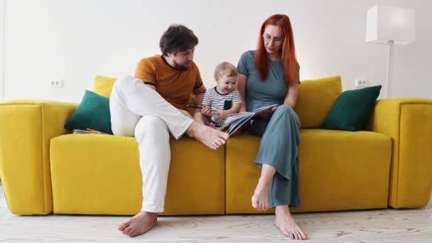 一家人坐在黄色的沙发上，一起看书- -婴儿把书吸引住了 — 图库视频影像