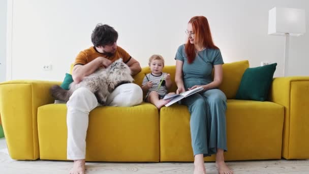 Familia sentada en un sofá amarillo con un gran gato esponjoso y el bebé dibuja en el libro — Vídeo de stock