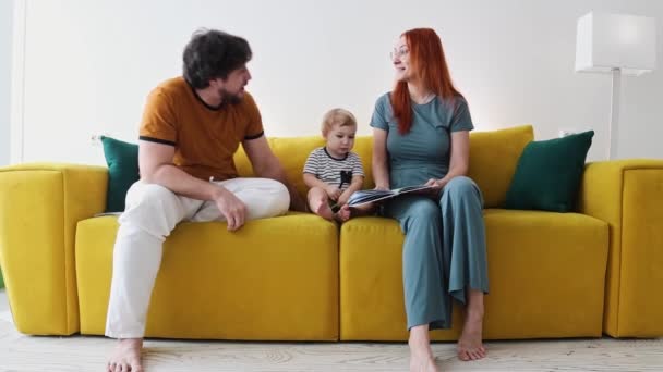 Keluarga duduk di sofa kuning dan membaca buku bersama-sama bayi memegang pena dan lily — Stok Video