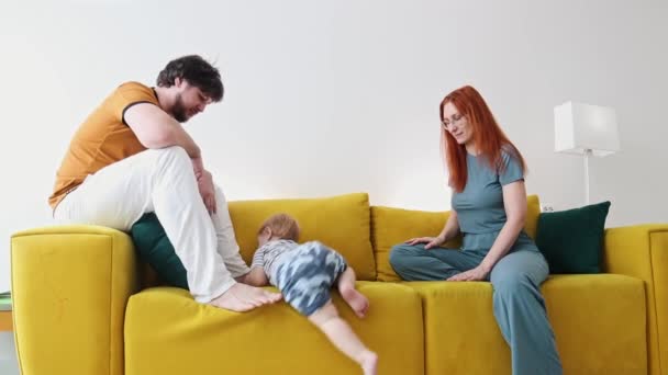 Família sentada no sofá amarelo e o bebê rastejando para baixo do sofá — Vídeo de Stock