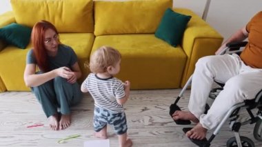 Küçük bir çocuk televizyondan uzaktan kumandayı monte eder. Annesinden babasına tekerlekli sandalyede yürür.