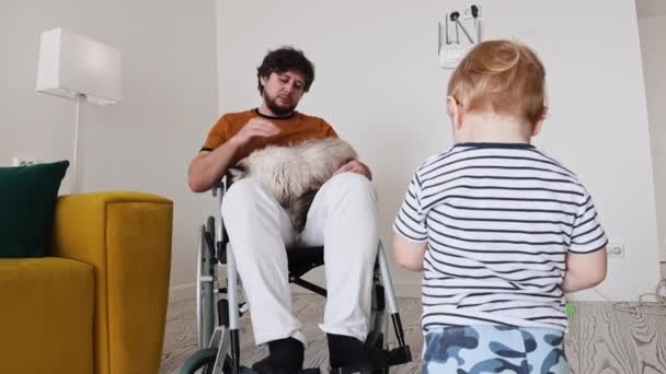 Padre en silla de ruedas con un gato esponjoso en sus piernas y su pequeño hijo jugando delante de él — Vídeo de stock
