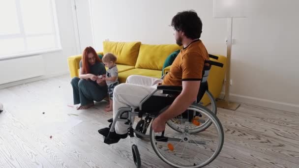 La mujer que juega con su hijo pequeño y el padre en silla de ruedas se acerca a ellos — Vídeo de stock