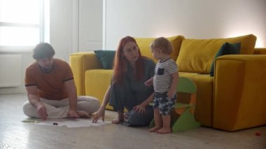 Aile yere oturur. Kızıl kadın kağıda resim çizer ve bebeği onu izler.
