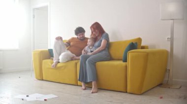 Küçük bebekli bir aile sarı kanepede oturup televizyon izliyor.