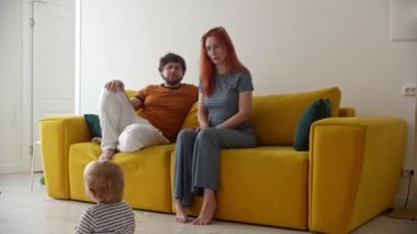 Genç ebeveynler sarı koltukta oturur ve küçük bebeklerinin odada yürümesini izlerler.