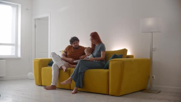A család ül a sárga kanapén, a baba rajzol valamit egy füzetbe ceruzával. — Stock videók