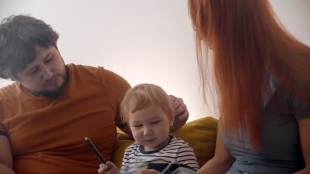 Aile kanepede oturuyor ve ebeveynler bebeklerine bir kitap gösteriyor. Bebek kalem çiğniyor. — Stok video