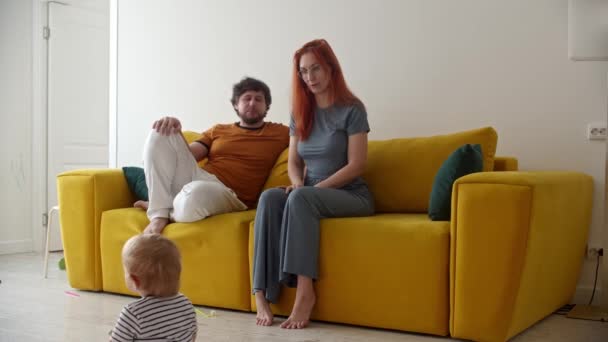 年轻的父母坐在黄色的沙发上看着他们的小宝宝在房间里走来走去 — 图库视频影像