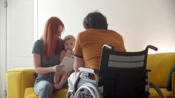 Mutter und Baby zeichnen in Notizbuch und Vater im Rollstuhl macht seinem Sohn lustige Gesichter — Stockvideo