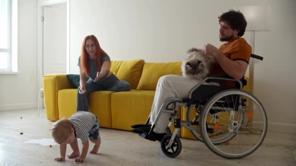 Mannen i rullstol tittar på sin fru och sitt barn leker med TV-fjärrkontrollen — Stockvideo