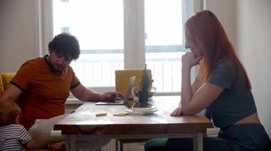 Aile rutini. Babası laptopla çalışıyor. Karısıyla masada oturuyor ve küçük oğlu masanın üstünde emekliyor.